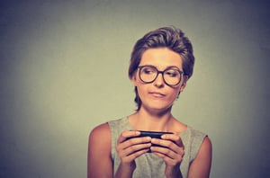 Portrait young angry woman with glasses unhappy, annoyed by something on cell phone while texting receiving bad sms text message isolated grey wall background. Human face expression emotion reaction